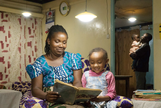 Where We Work Box - Credit - SHS_Ignite Rwanda-IRENA An adult and a child sit indoors reading a book together under electric lights in a home setting. In the background, another adult holds a young child, with household furnishings visible.
