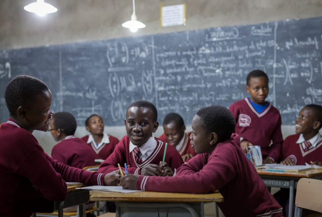 A group of children sit around a table in a classroom or training room, engaged in a discussion, with a chalkboard covered in writing visible behind them.