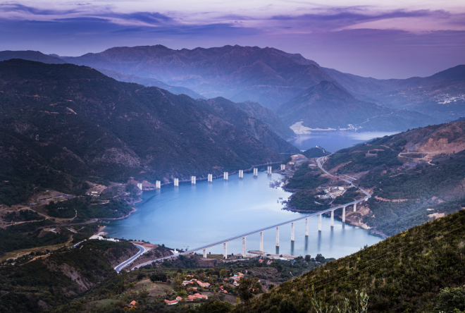 Credit: Noureddine-Belfethi_GettyImages Wide landscape view of a winding river and a hydropower plant surrounded by mountainous valley at dusk, with surrounding hills and lights from nearby settlements visible along the shoreline.