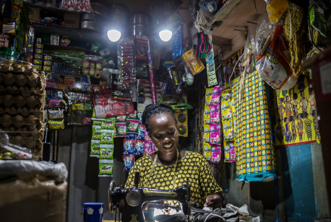 A woman works at a sewing machine inside her small, brightly lit shop.