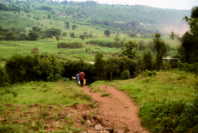 Two people walk on a dirt path through lush green countryside with distant hills
