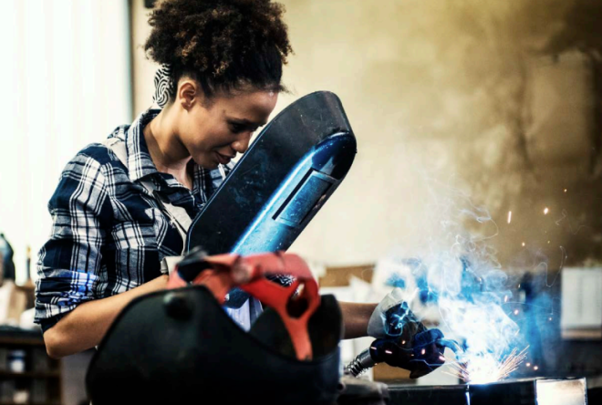 Person using welding equipment on a metal workpiece, with bright welding sparks, protective gear, and workshop tools visible in an industrial setting.