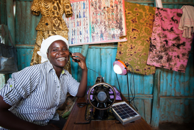 Energy Access - Solar Aid Woman seated indoors using a sewing machine, working with colorful fabric in a small workshop or market setting.
