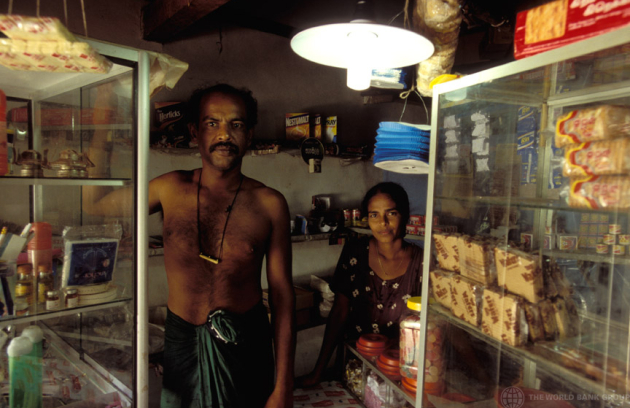 A small shop interior lit by electric lights, with shelves stocked with packaged goods and beverages. Two people stands behind a glass display counter, with products visible on both sides of a narrow aisle.