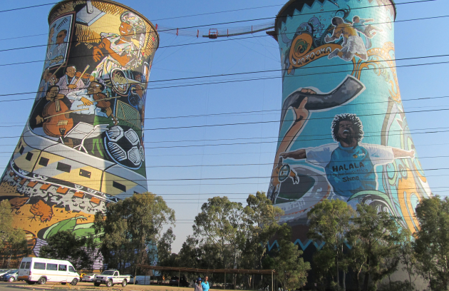 Two large cylindrical coal towers painted with colorful murals, connected by cables overhead, with trees and a street visible below under a clear sky.