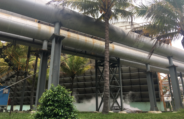 Industrial pipes and infrastructure run above ground among greenery, with metal structures and connected piping visible outdoors at an energy or utility facility.