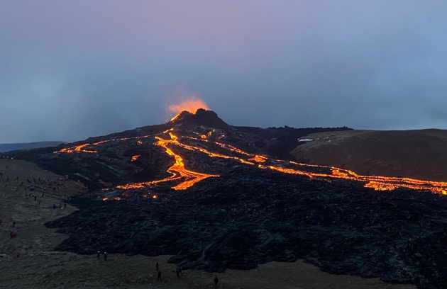 A volcanic landscape showing lava flowing across dark ground at night or dusk, with glowing streams of molten rock visible against the surrounding terrain.
