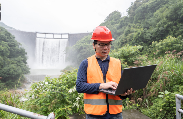 A field worker wearing a hard hat and high‑visibility vest uses a laptop at an outdoor site, with a hydropower dam and flowing water visible in the background.