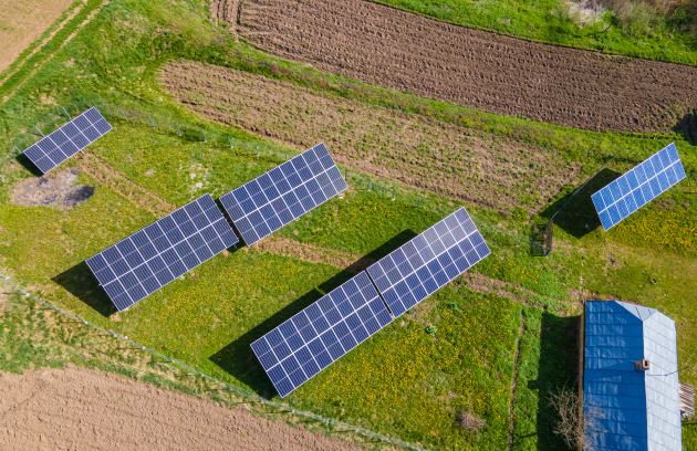 Aerial view of a small solar panel installation arranged in rows on a grassy field, with nearby vegetation and a small structure visible at the edge of the site.