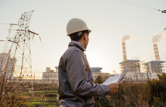 Industrial worker wearing a hard hat and grey work jacket while reviewing documents near a power plant