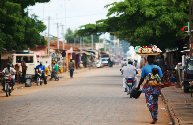 Outdoor street scene with trees, pedestrians, and vehicles lining the sandy road