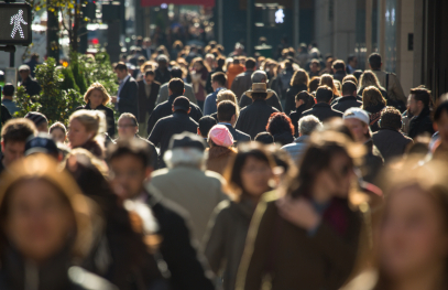A dense crowd of people walks along a city street, with individuals wearing coats and backpacks, suggesting a busy urban environment during commuting hours.