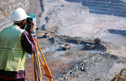 A worker wearing a safety helmet and high‑visibility vest stands at an elevated viewpoint, surveying a large open industrial site with heavy machinery, earthworks, and infrastructure spread across a rocky landscape.