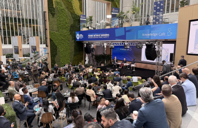 Large audience seated at an indoor public event with panelists speaking on a stage, surrounded by banners and screens labeled “Knowledge Café,” with cameras and event staff visible.