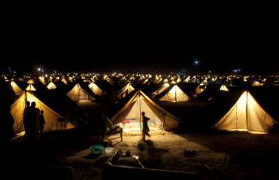 People gathered inside a row of illuminated tents at night, with warm lights creating a shared outdoor setting.