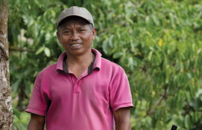 Person standing outdoors beside greenery, wearing a cap and a short‑sleeved shirt, with trees and foliage in the background.