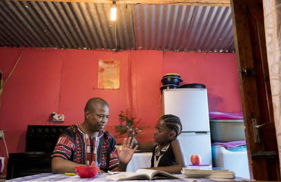 A father helps his daughter with her homework while sitting at a table inside a modest home.