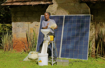A woman operates a small milling machine powered by solar panels outside a rural building.