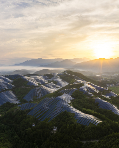 Aerial view of a large solar power installation covering rolling hills at sunrise, with rows of solar panels set among green vegetation and mountains in the distance.