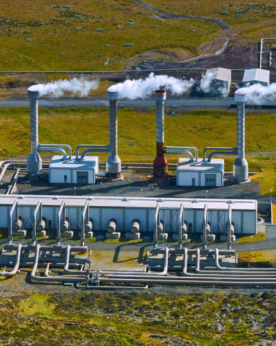 PDF p. 21 report Geothermal Energy: Unveiling the Socioeconomic Benefits Aerial view of an energy facility with multiple industrial buildings, pipes, and tall vents releasing steam, set in an open landscape.