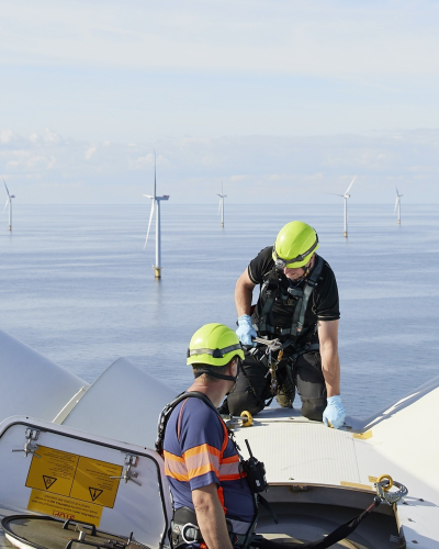 Technicians work atop an offshore wind turbine, with wind turbines visible across the sea.