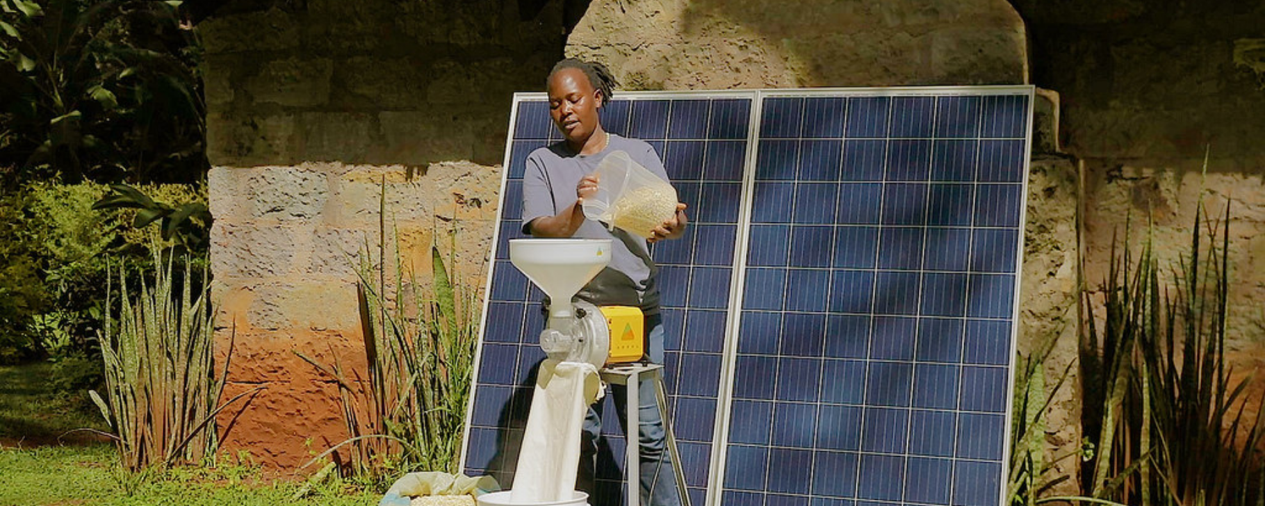 Electricity Access Banner A person operates a grain milling machine powered by solar panels installed outdoors, with two large solar panels positioned behind the equipment.