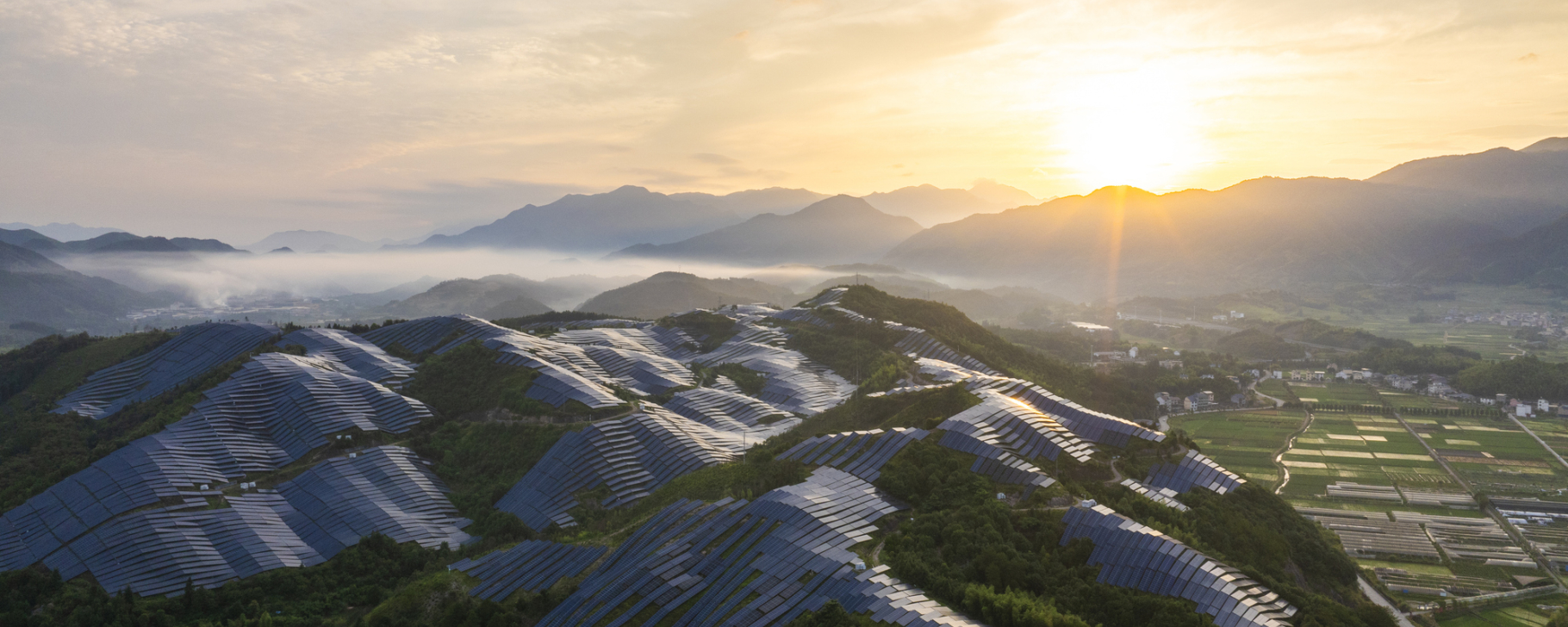 Aerial view of a large solar power installation covering rolling hills at sunrise, with rows of solar panels set among green vegetation and mountains in the distance.