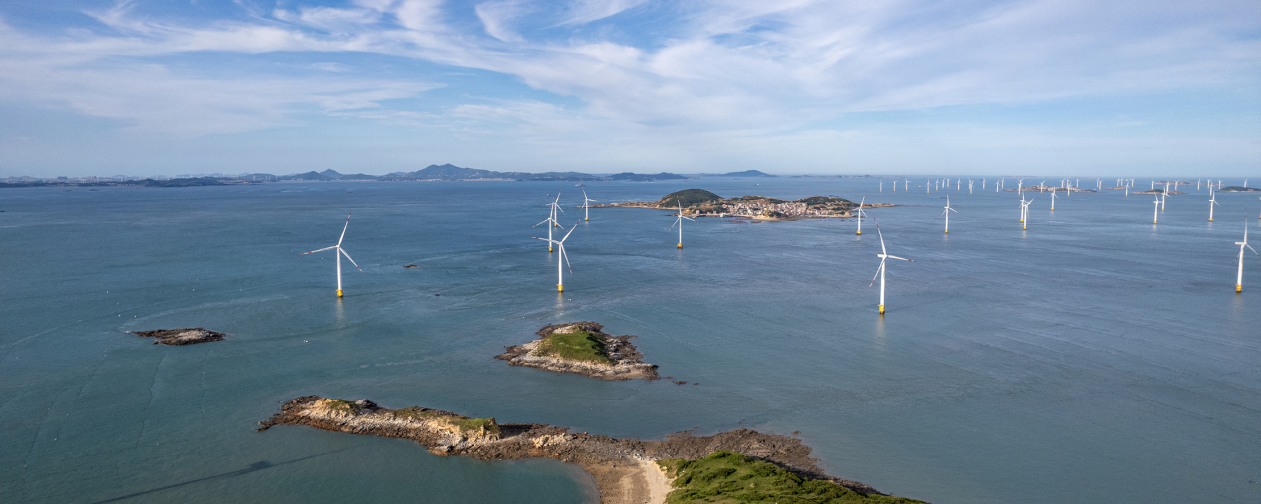 Energy Solutions - Banner A coastal landscape with offshore wind turbines installed in the water near a shoreline, with bridges and infrastructure visible in the background.