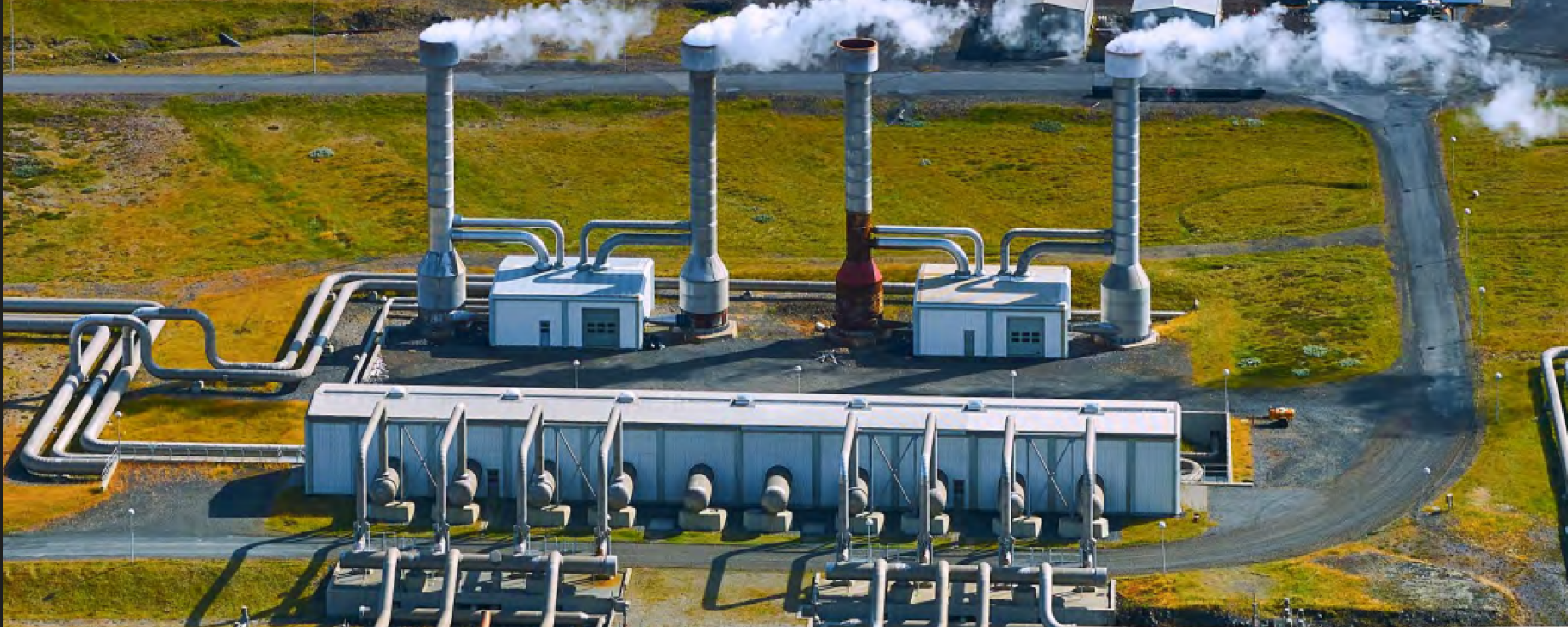 PDF p. 21 report Geothermal Energy: Unveiling the Socioeconomic Benefits Aerial view of an energy facility with multiple industrial buildings, pipes, and tall vents releasing steam, set in an open landscape.
