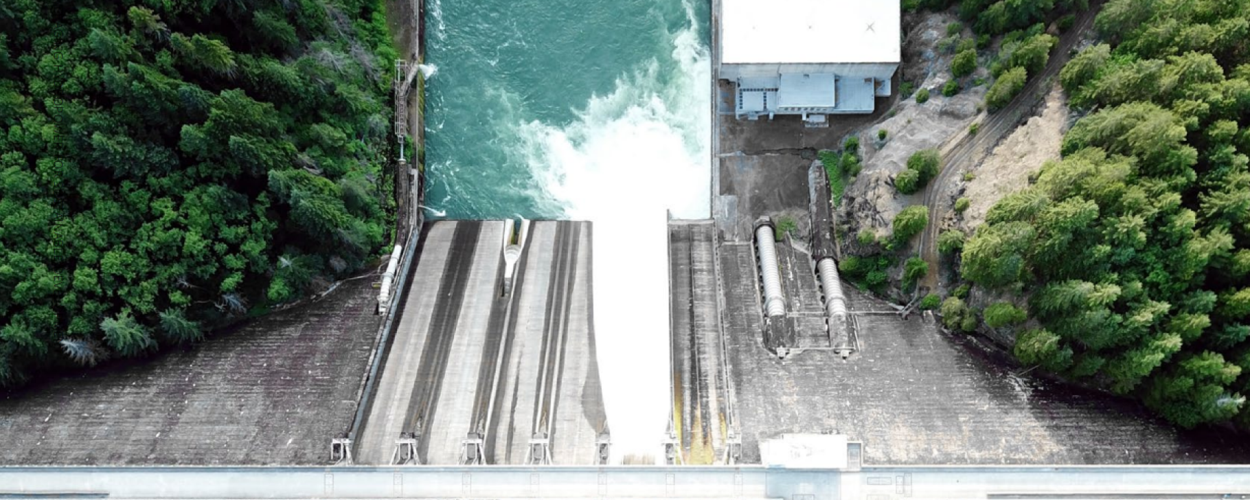 Image source: P. 10 report “Hydropower: Unveiling the Socioeconomic Benefits” Overhead view of a hydropower dam with water flowing through spillways into a river below, surrounded by forested terrain and operational infrastructure.