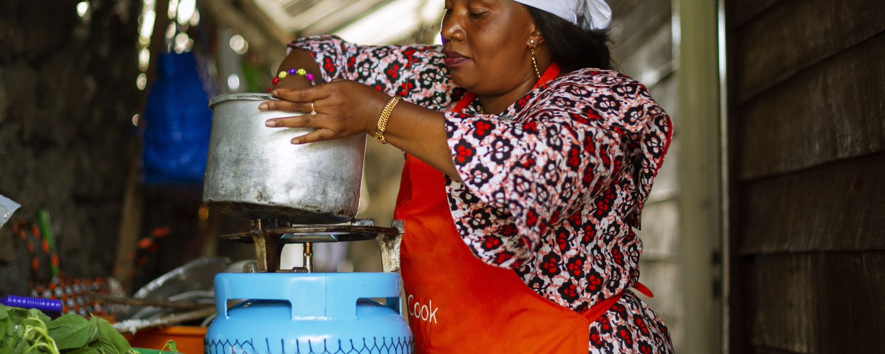 Person cooking indoors using a gas stove, lifting a metal pot while wearing a head covering and apron, with food ingredients and cooking utensils nearby.