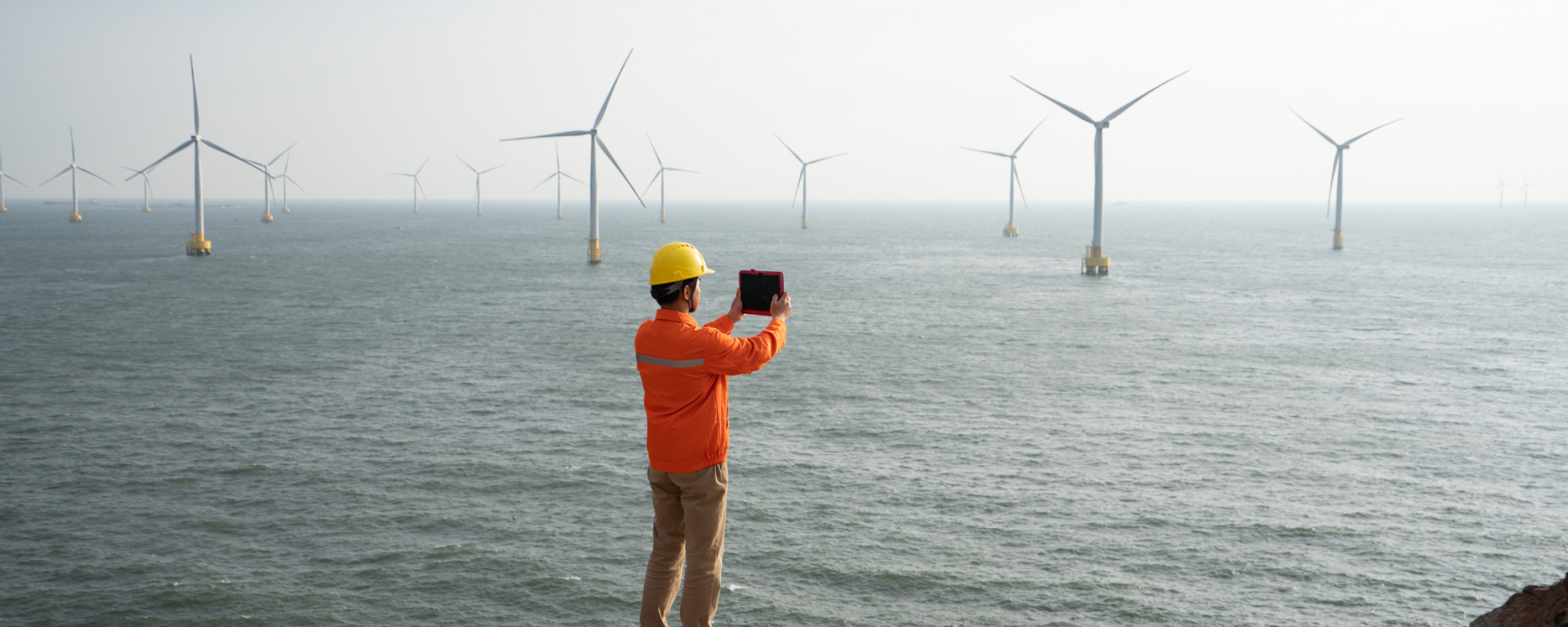 Energy Transition Banner Person standing on a rocky outcrop overlooking the ocean, with offshore wind turbines visible in the distance.