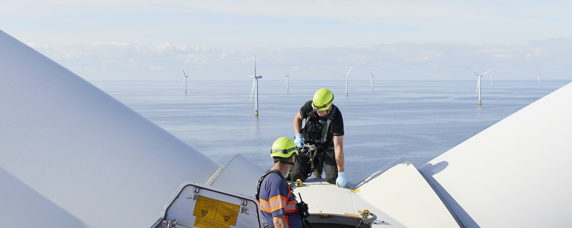 Technicians work atop an offshore wind turbine, with wind turbines visible across the sea.