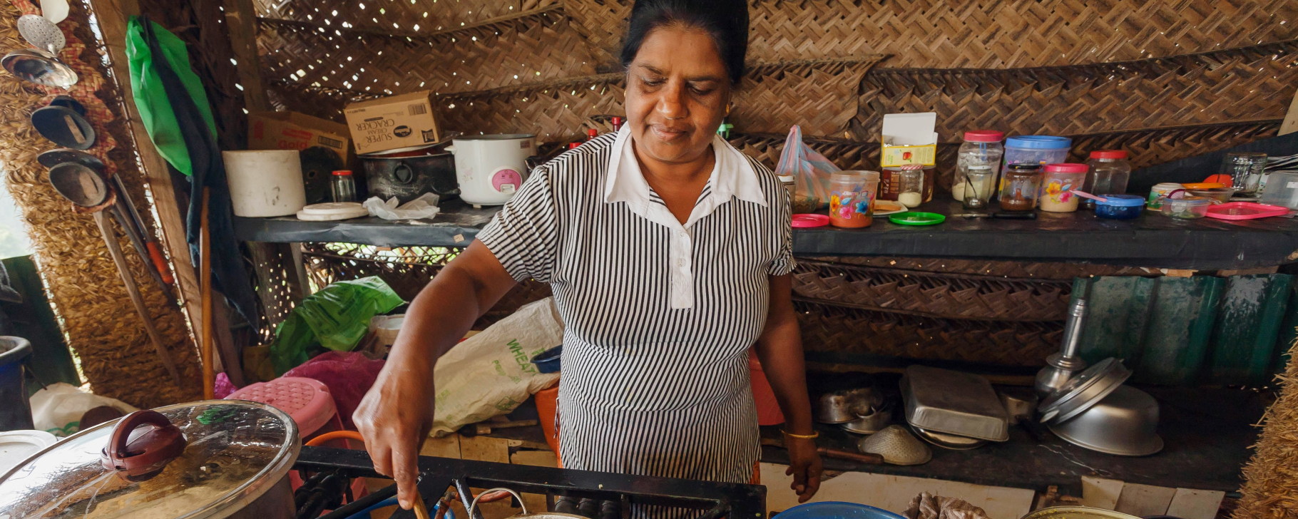 Person stirring food in a pot on a stove inside a woven-wall kitchen with cooking utensils.