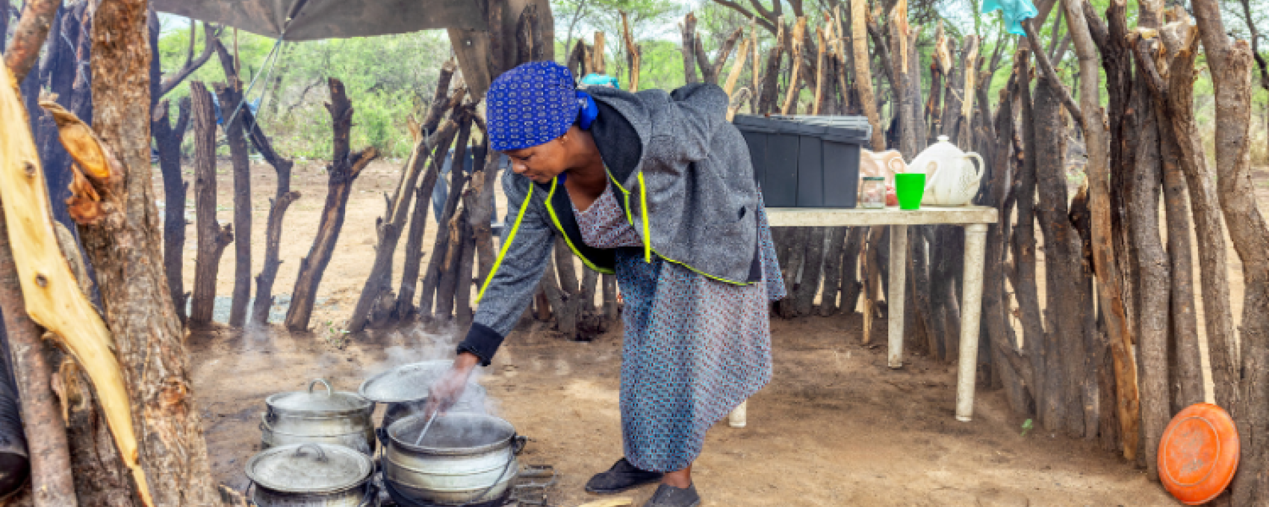 A person cooking outdoors over an open wood fire, stirring food in several metal pots beneath a makeshift shelter surrounded by trees and wooden fencing.