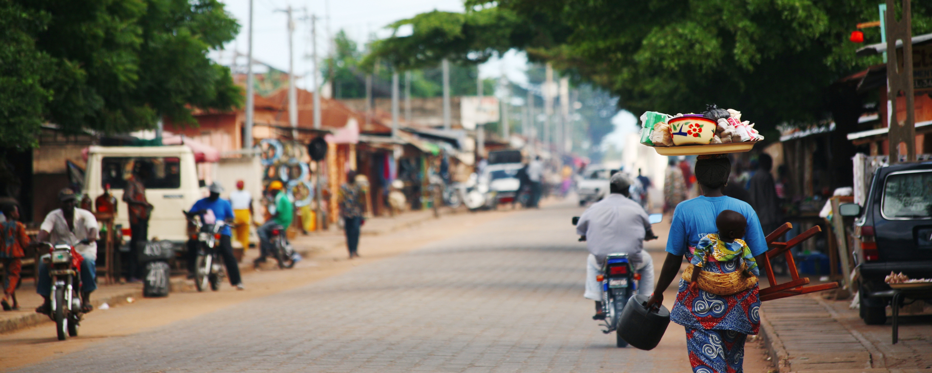 Outdoor street scene with trees, pedestrians, and vehicles lining the sandy road