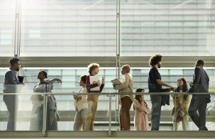 People standing and talking on an indoor elevated walkway with glass railings, holding notebooks and tablets inside a modern office building filled with natural light.