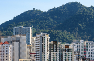 Urban residential buildings clustered at the base of a forested hillside, with mid‑rise and high‑rise apartments against a clear blue sky.