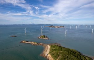 Aerial view of offshore wind turbines positioned in the sea near small rocky islands, with a distant coastline under a partly cloudy sky.