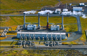 Aerial view of an energy facility with multiple industrial buildings, pipes, and tall vents releasing steam, set in an open landscape.
