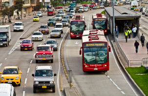 Busy urban roadway with red bus rapid transit vehicles traveling in dedicated lanes alongside mixed traffic, with a covered bus station and pedestrians on the sidewalk.
