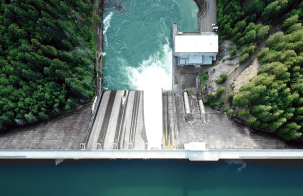 Overhead view of a hydropower dam with water flowing through spillways into a river below, surrounded by forested terrain and operational infrastructure.
