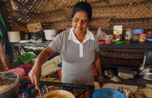 Person stirring food in a pot on a stove inside a woven-wall kitchen with cooking utensils.