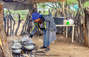 A person cooking outdoors over an open wood fire, stirring food in several metal pots beneath a makeshift shelter surrounded by trees and wooden fencing.