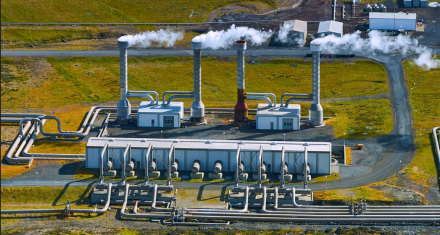 PDF p. 21 report Geothermal Energy: Unveiling the Socioeconomic Benefits Aerial view of an energy facility with multiple industrial buildings, pipes, and tall vents releasing steam, set in an open landscape.