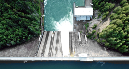 Image source: P. 10 report “Hydropower: Unveiling the Socioeconomic Benefits” Overhead view of a hydropower dam with water flowing through spillways into a river below, surrounded by forested terrain and operational infrastructure.