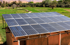 Solar panels mounted on the roof of a small building in a rural setting, with trees and open land visible around the structure.