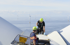Technicians work atop an offshore wind turbine, with wind turbines visible across the sea.