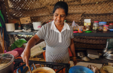 Person stirring food in a pot on a stove inside a woven-wall kitchen with cooking utensils.