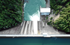 Overhead view of a hydropower dam with water flowing through spillways into a river below, surrounded by forested terrain and operational infrastructure.